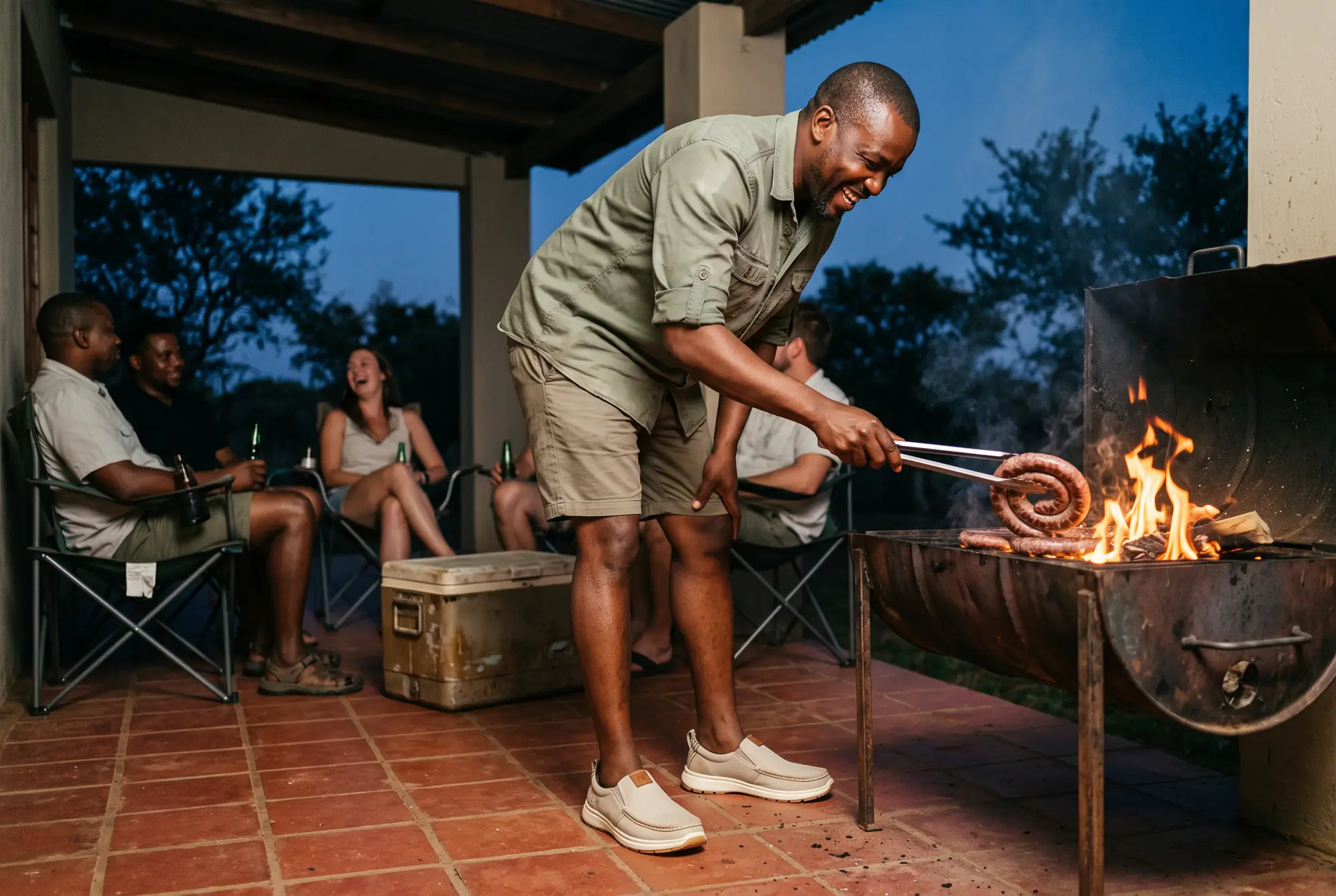 A laughing man turns boerewors on a fire on a farm stoep at dusk while friends sit behind him, wearing stone R01s