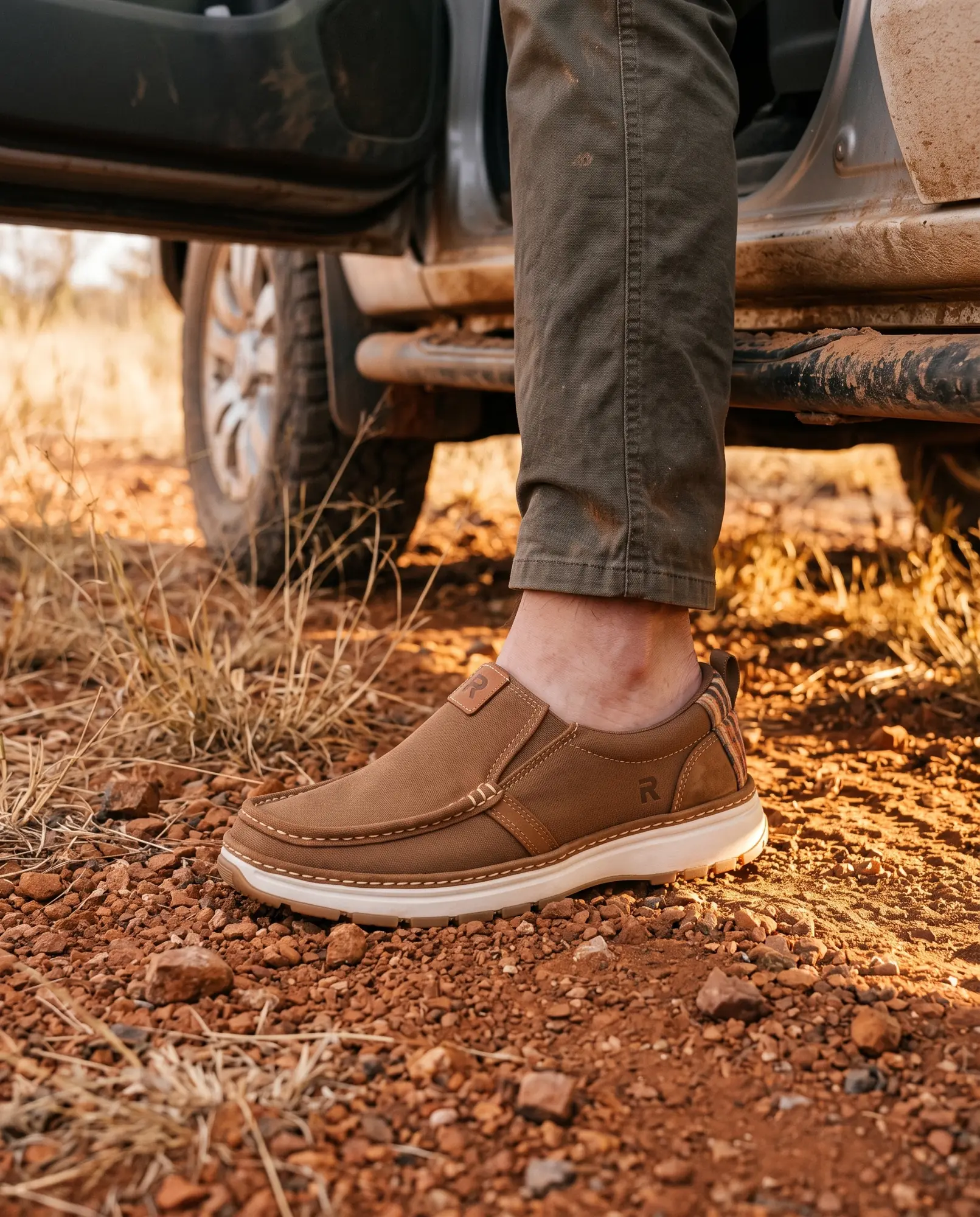 A foot stepping hands-free into a tobacco R01 slip-on on red gravel beside a 4x4