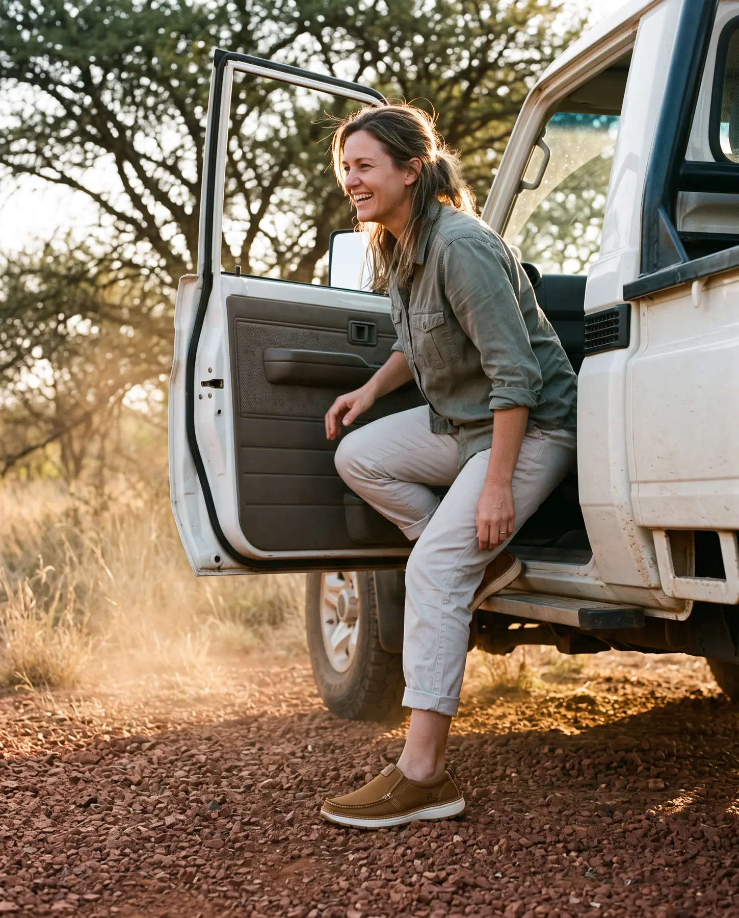 A laughing woman steps down from the door of a dusty white bakkie onto red gravel, wearing tobacco R01W slip-ons