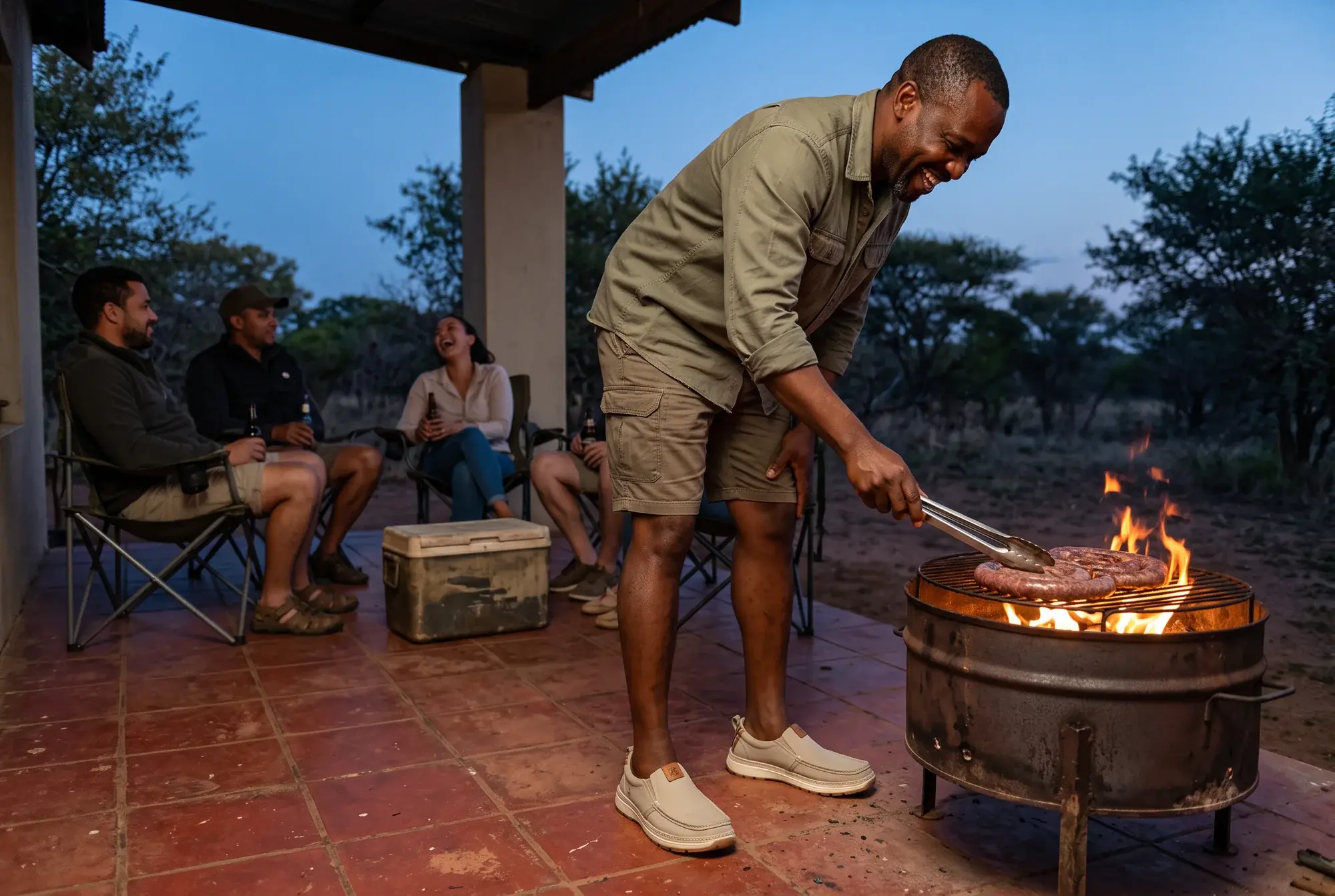 A laughing man turns boerewors on a fire on a farm stoep at dusk while friends sit behind him, wearing stone R01s