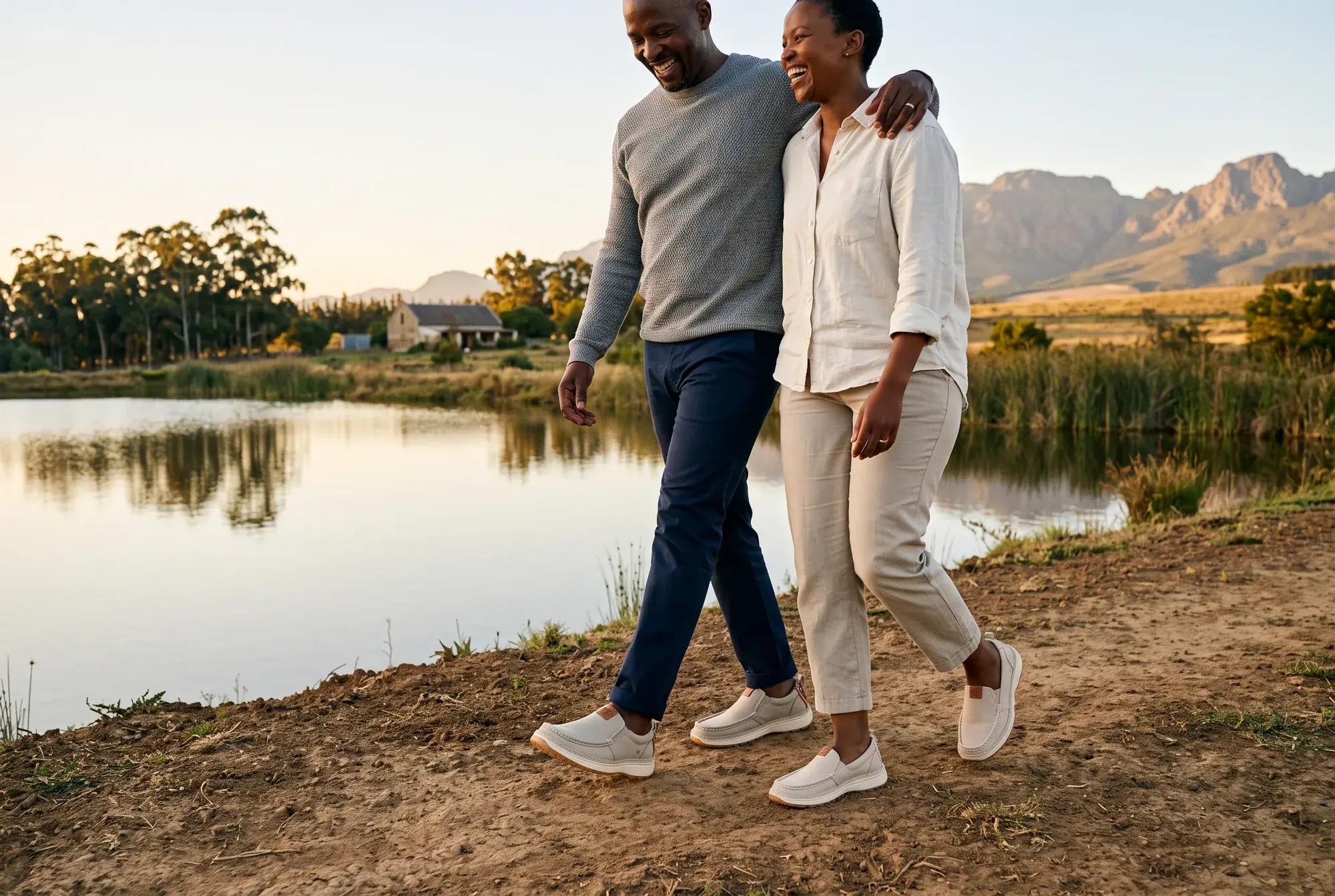 A laughing couple walks along a farm dam at golden hour below the mountains, both wearing stone slip-ons, his R01 and her R01W