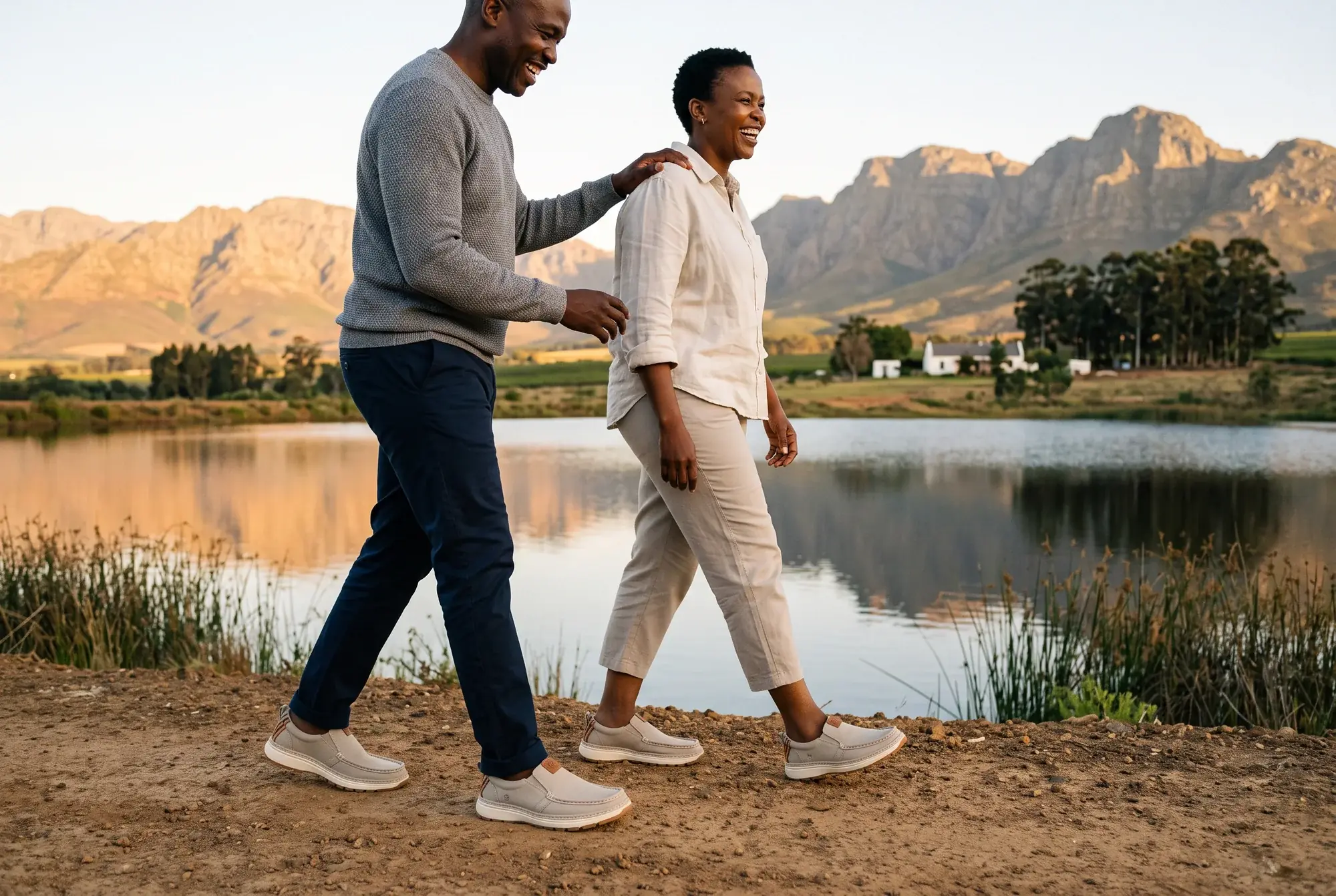 A laughing couple walks along a farm dam wall at golden hour below the mountains, both wearing stone slip-ons, his R01 and her R01W
