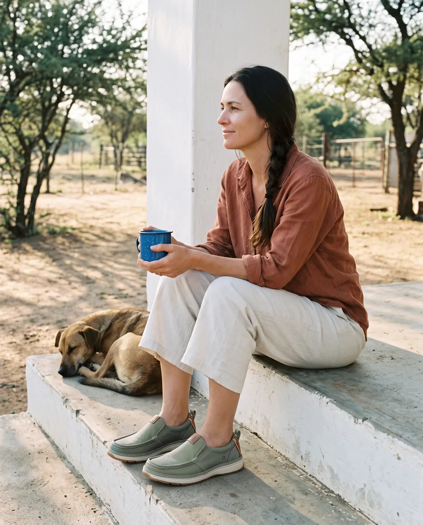 A woman with a long dark plait sits on whitewashed farmhouse steps with a coffee mug, wearing sage R01W slip-ons, a dog asleep beside her