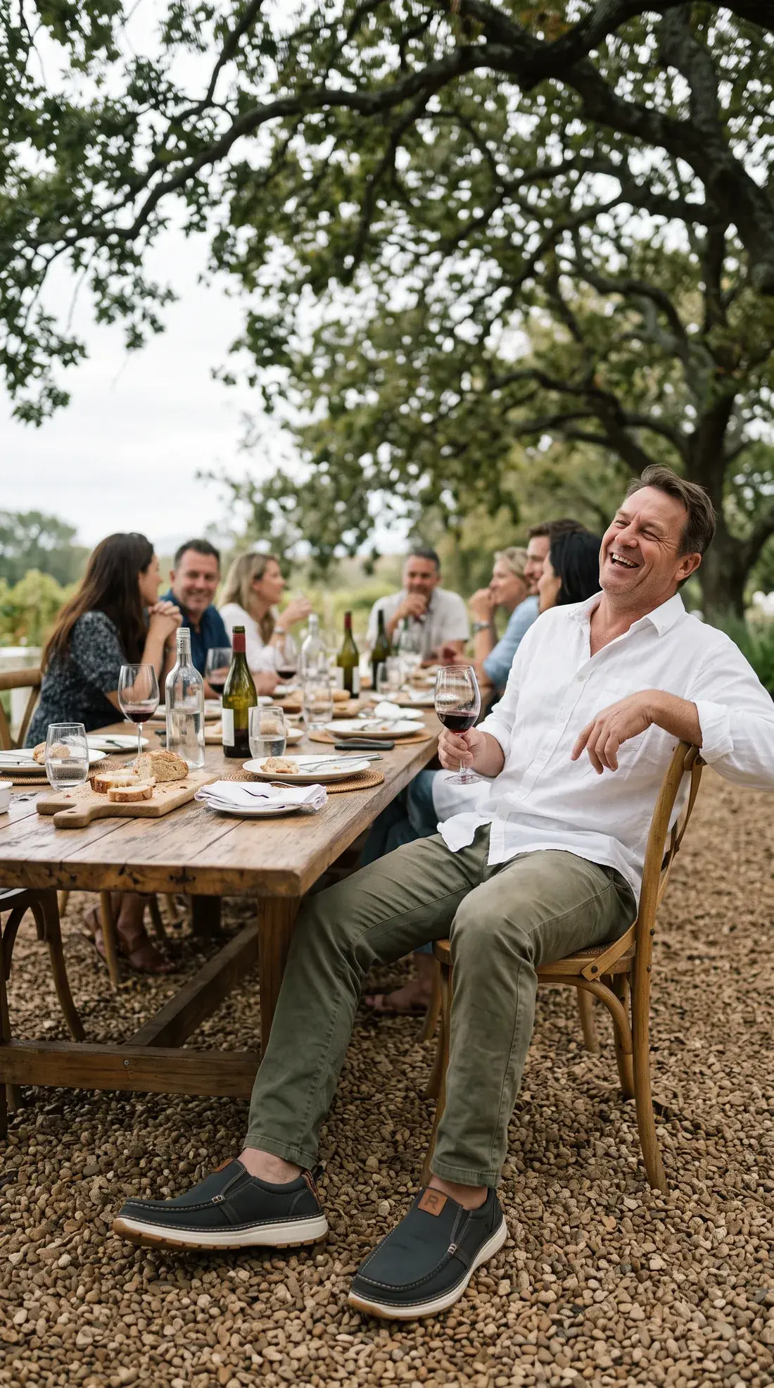 A man leans back laughing at the end of a long lunch table under oak trees on a Cape wine estate, glass of red in hand, wearing charcoal R01 slip-ons