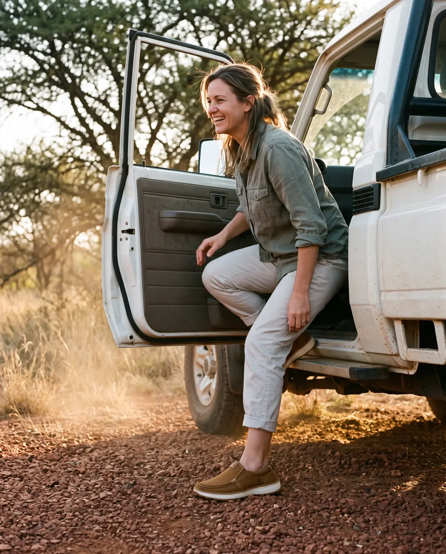 A laughing woman steps down from the door of a dusty white bakkie onto red gravel, wearing tobacco R01W slip-ons