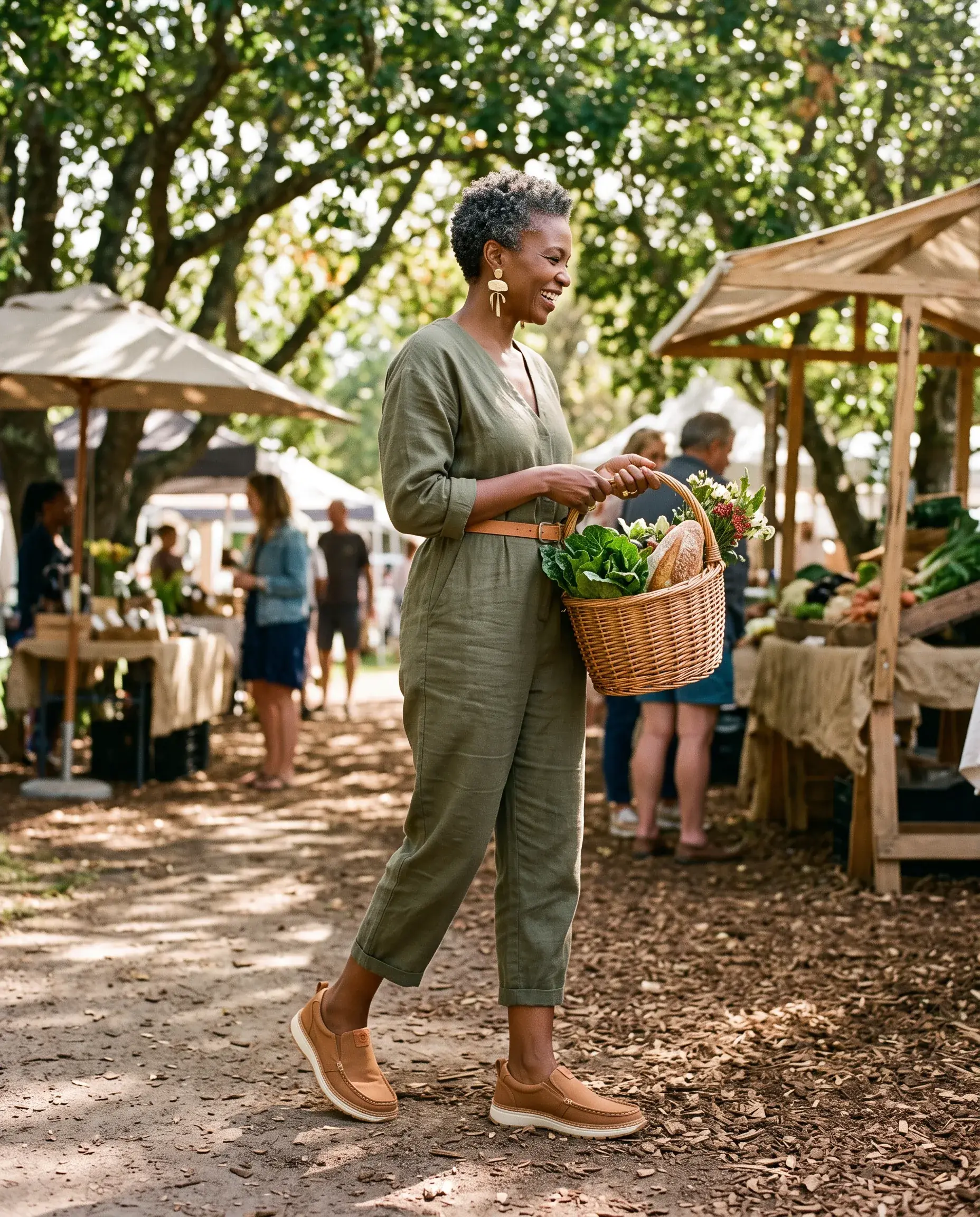 A woman with a basket of vegetables walks through a shaded farmers' market, wearing tobacco R01W slip-ons
